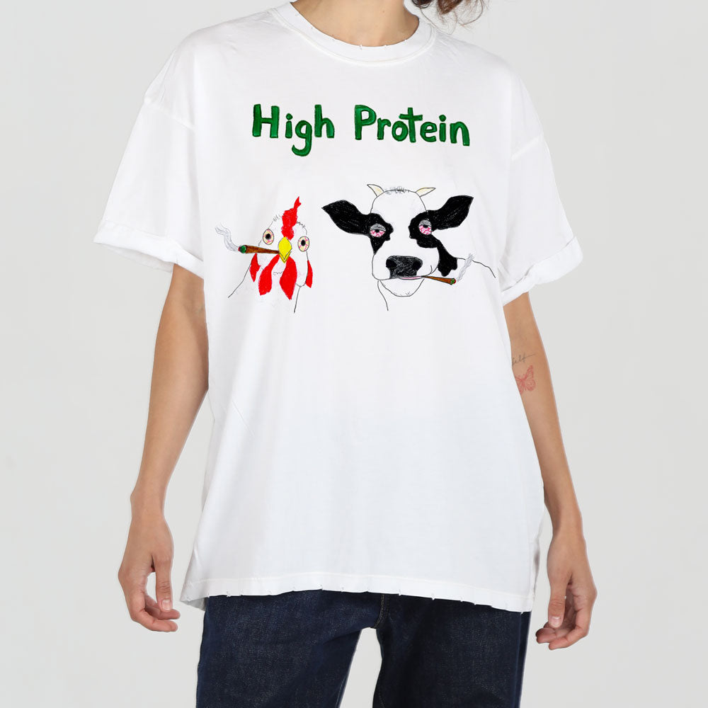 A person wearing a white t-shirt with a humorous design featuring a cow and a chicken, both holding forks, titled "high protein.