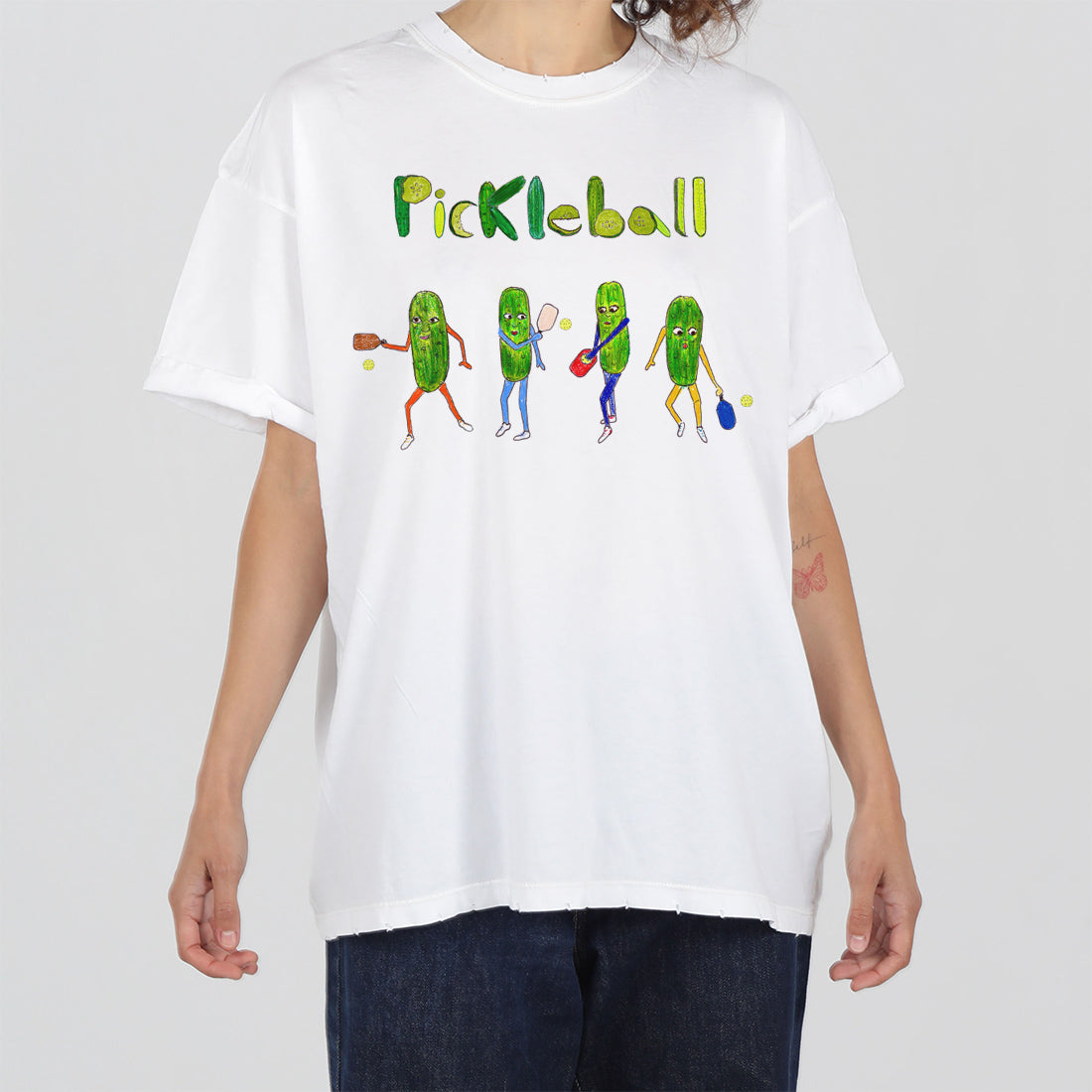 A person wearing a white t-shirt with a colorful illustration of anthropomorphic pickles playing pickleball. the word "pickleball" is printed above in green.