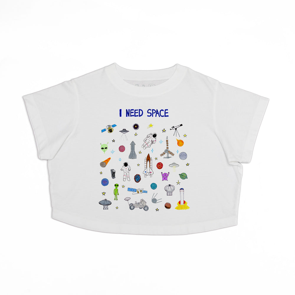 A white crop top t-shirt with the phrase "i need space" in blue, surrounded by colorful, whimsical drawings of space-themed illustrations like rockets, aliens, and planets.