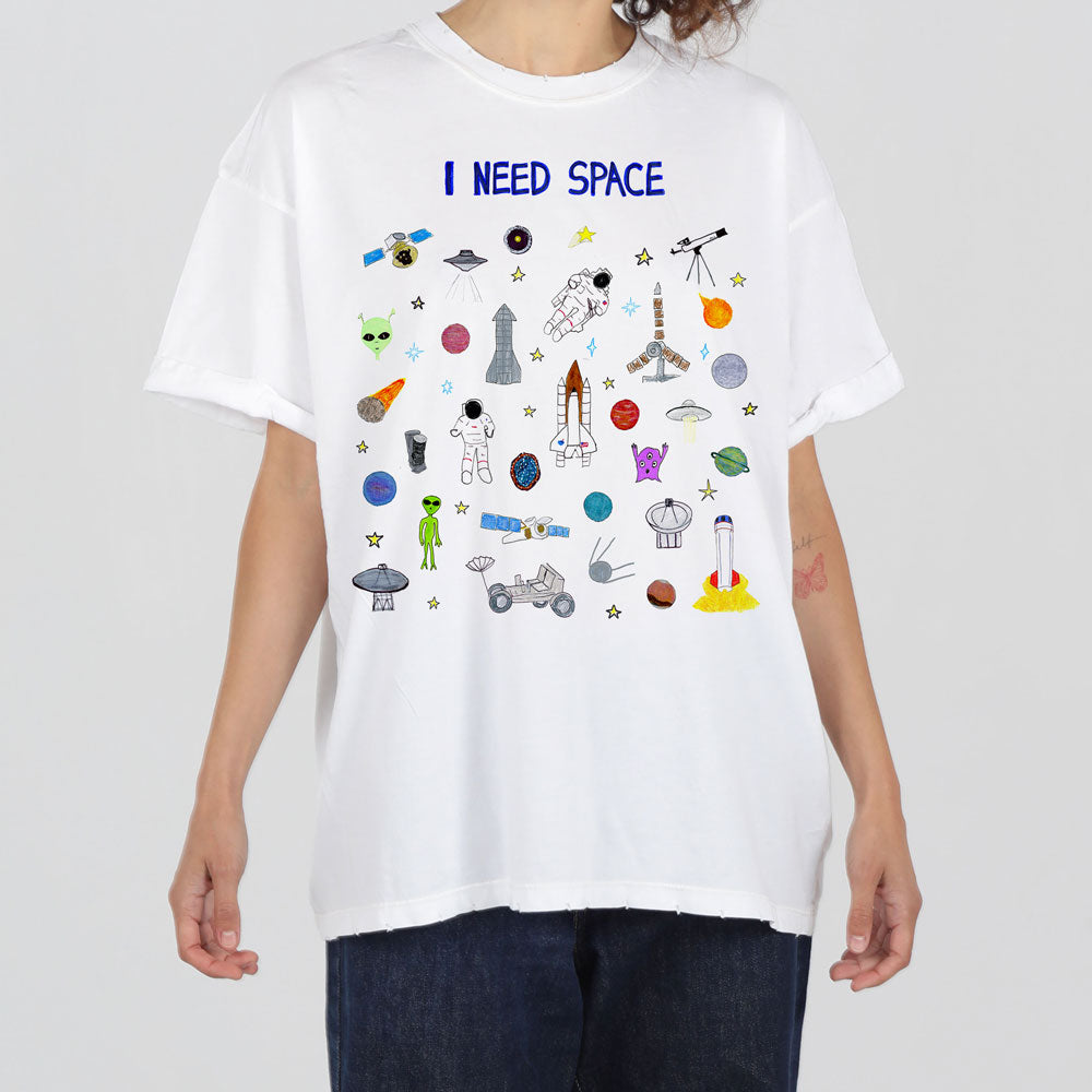 A person wearing a white t-shirt with various colorful space-themed graphics and the text "i need space" at the top. the designs include aliens, rockets, planets, and telescopes.