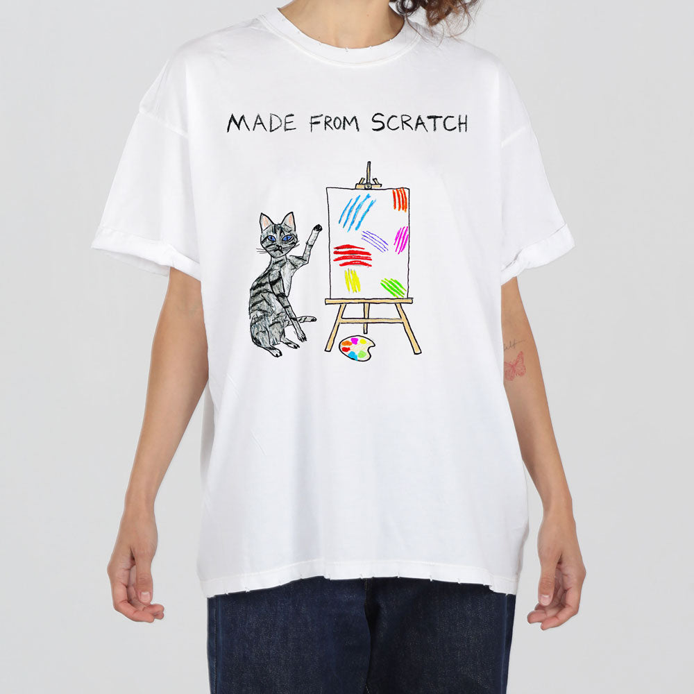A person in a white t-shirt featuring a printed design of a cat beside an easel with colorful strokes and a paint palette.