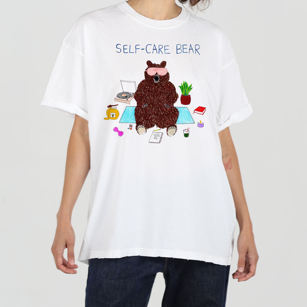 A person wearing a white t-shirt featuring a graphic of a brown bear with sunglasses, surrounded by self-care items like tea and plants, with the caption "self-care bear".