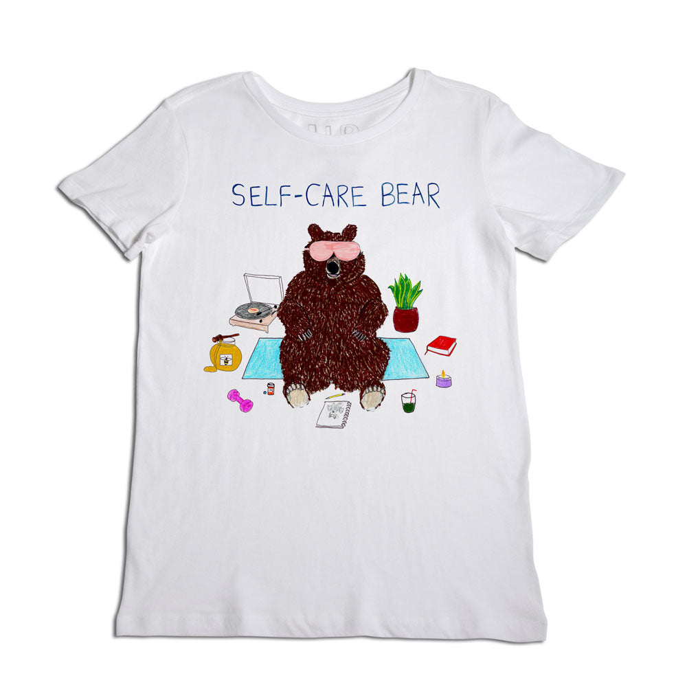 White t-shirt with a graphic of a brown bear labeled "self-care bear," sitting with a laptop, books, a coffee mug, and plants, promoting self-care and relaxation.