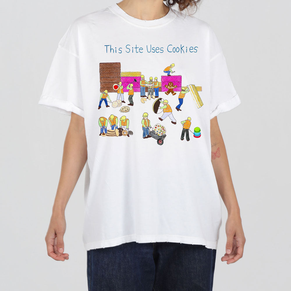A person wearing a white t-shirt with a colorful graphic of cartoon characters resembling construction workers and the text "this site uses cookies".
