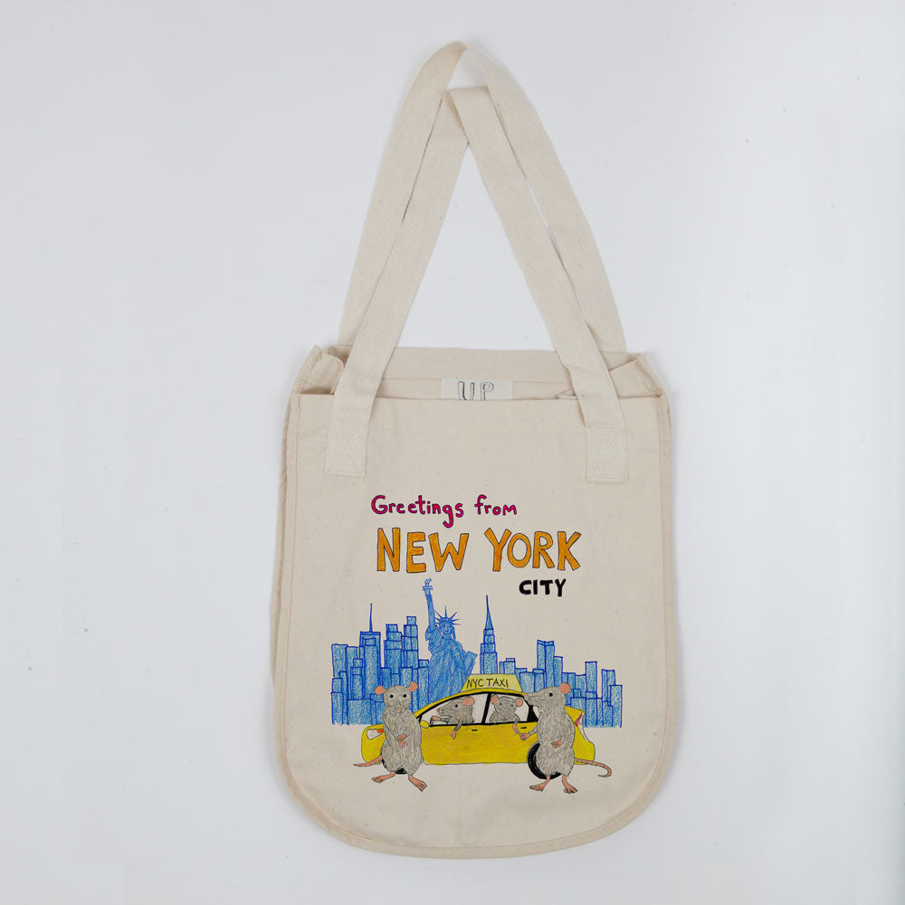 Beige tote bag with "greetings from new york city" text and an illustration featuring the nyc skyline and two rats carrying a yellow taxi. the bag has a long strap and is displayed on a white background.