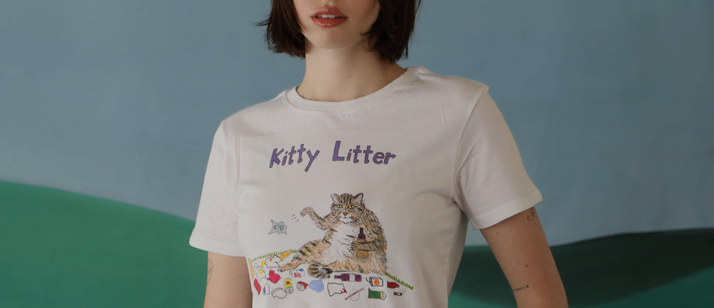 A white model with a blunt black bob stands with her hand on hips. She wears a graphic illustration tee that says "Kitty Litter" and shows a cat surrounded by trash. 