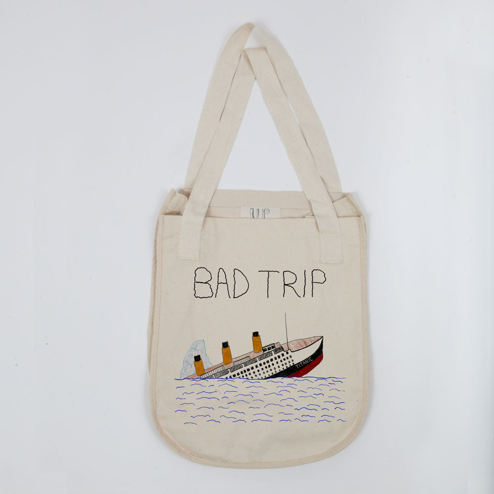 A cream-colored tote bag with the phrase "bad trip" printed above an illustration of a sinking ship surrounded by blue waves, displayed on a plain white background.