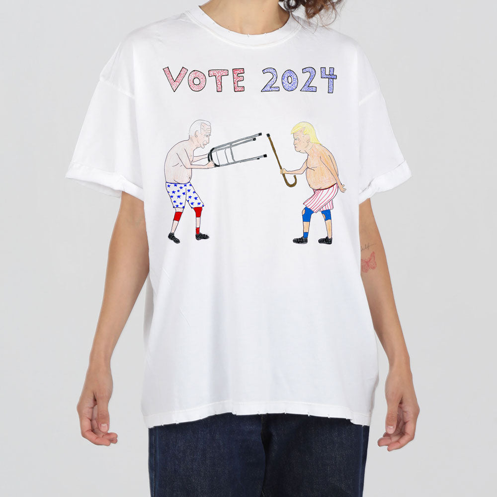 A person wearing a white t-shirt with "vote 2024" in pink letters and graphics of two people using a giant lipstick as a bazooka, under a bright studio light.