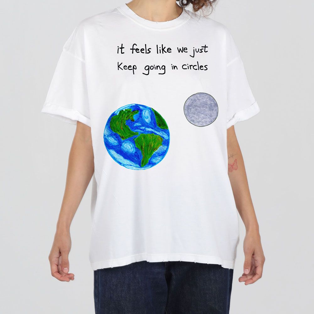 A person wearing a white t-shirt with a print of earth and the moon, and text saying "it feels like we just keep going in circles".
