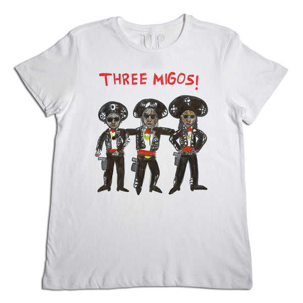 A white t-shirt featuring an illustration of three stylized characters dressed as "three amigos" with sombreros and colorful costumes. above the characters, text reads "three migos!" in red.