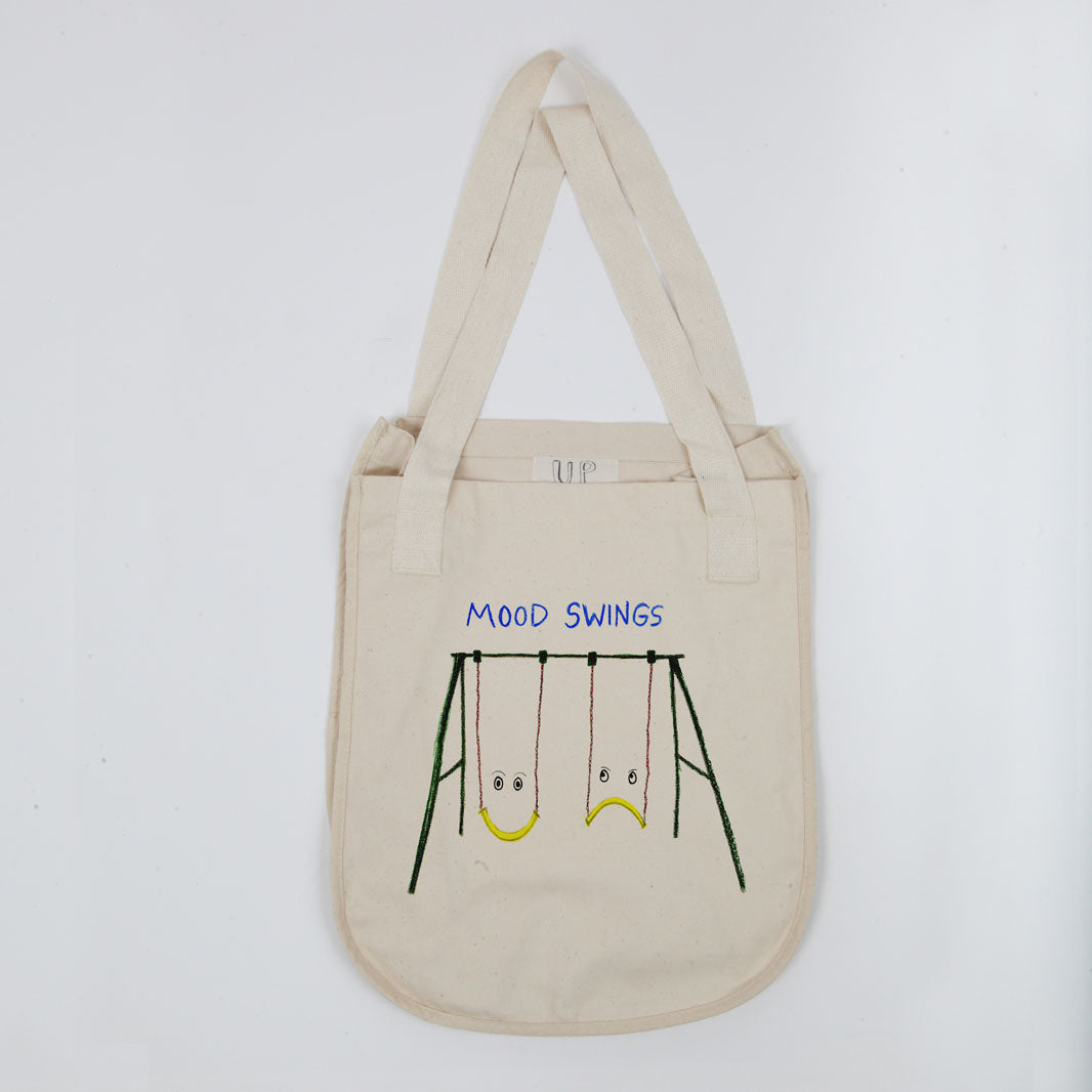 A beige canvas tote bag with a graphic depicting a swing set, labeled "mood swings" with two swings, one frowning and one smiling, against a plain background.