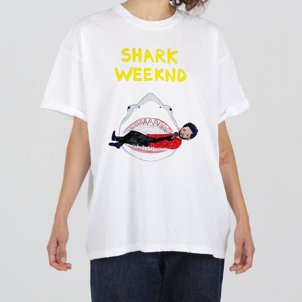 A person wearing a white t-shirt with a graphic design featuring a shark and the text "shark weekend" above it. the shark is depicted with an open mouth, inside which there is an illustration of a person.